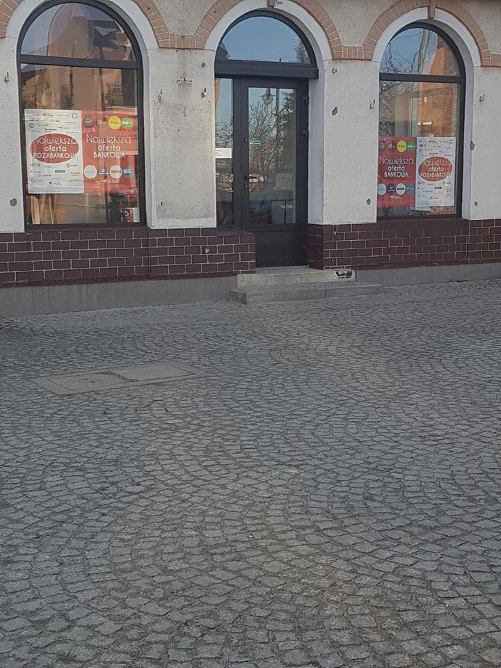 Fasada budynku z dwoma zaokrąglonymi oknami, na których widoczne plakaty z ofertą bankową, oraz ciemnymi drzwiami wejściowymi, całość na tle brukowanej ulicy.
