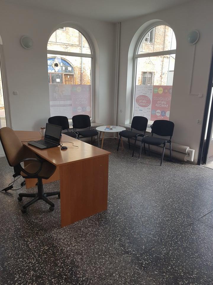 Biuro z biurkiem, laptopem i krzesłami w jasnym pomieszczeniu z dużymi oknami łukowymi i plakatami.