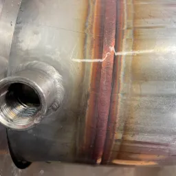 Spoina na metalowej rurze z widocznymi przebarwieniami termicznymi, z bliska widoczna drobna wada spawalnicza.