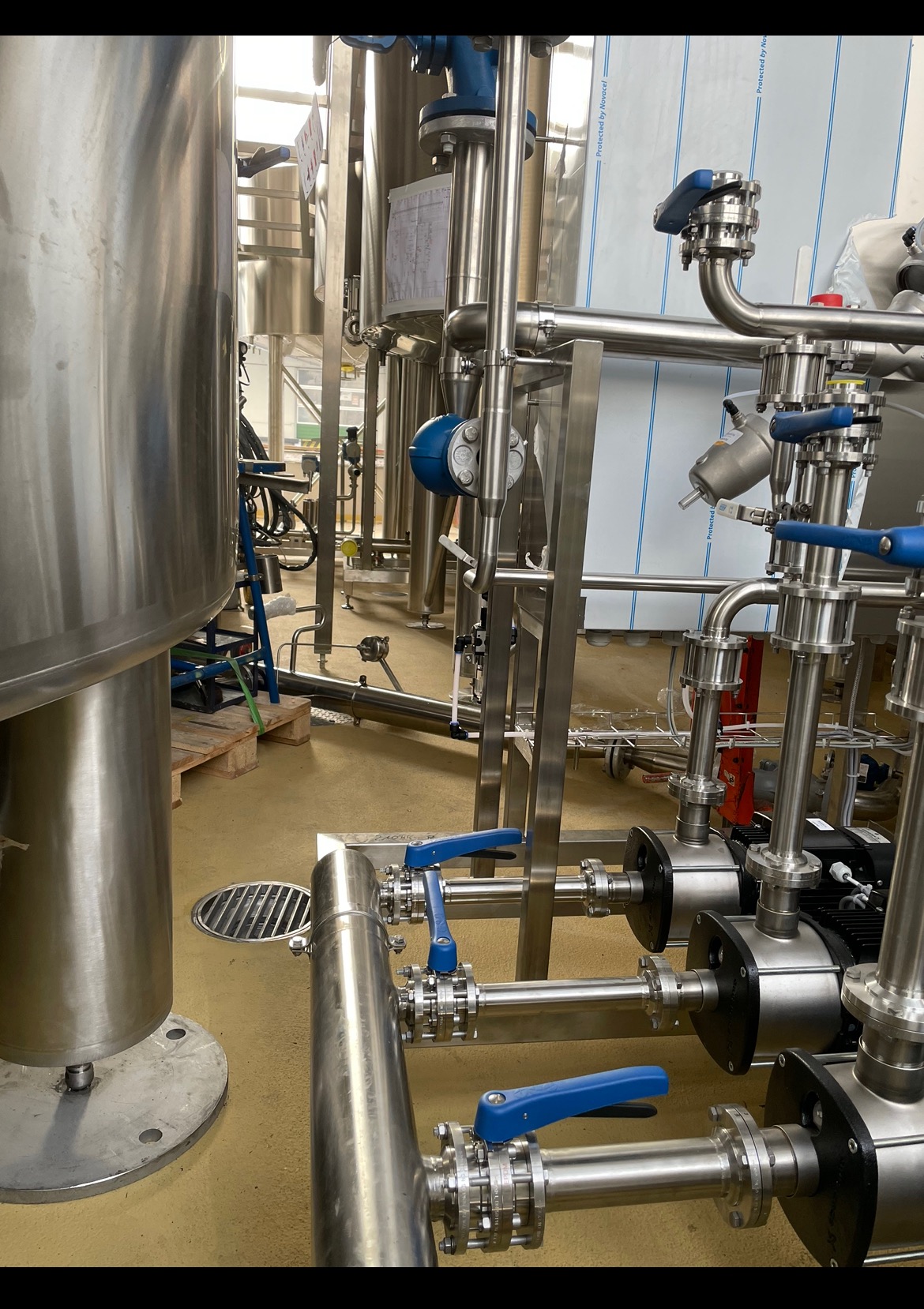 Instalacja przemysłowa z rurami ze stali nierdzewnej, zaworami z niebieskimi uchwytami i pompami, widoczne elementy konstrukcyjne i podłoga przemysłowa.
