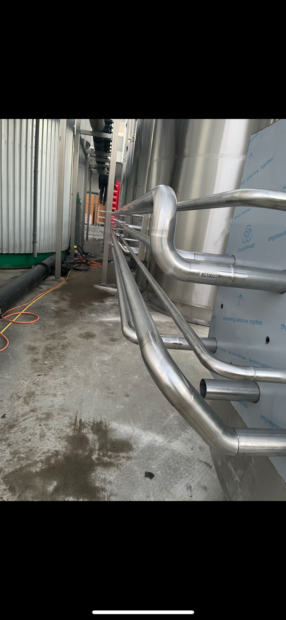 Połączenia spawane rur ze stali nierdzewnej na tle zbiorników przemysłowych, widok z dołu, widoczne ślady użytkowania na posadzce.