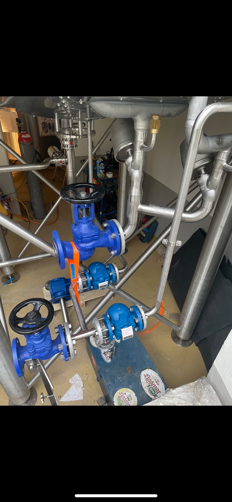 Złożona instalacja rurociągów ze stali nierdzewnej z niebieskimi zaworami kulowymi, częściowo zamocowana pomarańczowymi pasami transportowymi, z widoczną skrzynką narzędziową i butlą gazową w tle.