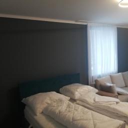 Daw-Bud Dawid Koziarz - Pokój po odświeżeniu: ciemnoszara ściana z białą listwą przypodłogową, łóżko z zielonym zagłówkiem i białą pościelą w folii, beżowa sofa z poduszkami w tle, białe zasłony w oknie.