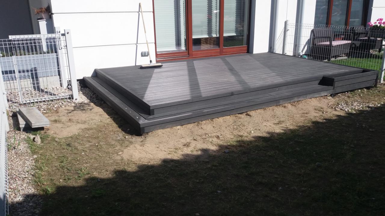 Nowoczesny, szary taras kompozytowy z dwoma stopniami, przylegający do białej ściany budynku, z widoczną szczotką opartą o ścianę, otoczony ziemią i niskim ogrodzeniem.