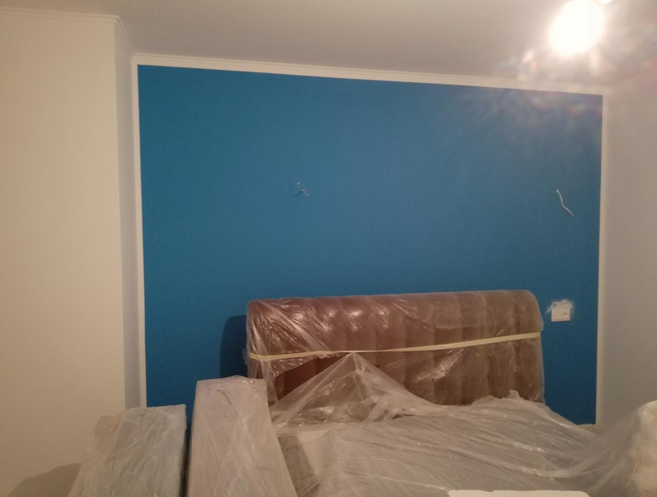 Świeżo pomalowana na niebiesko ściana w pokoju, zabezpieczone folią meble i materiały budowlane, biały sufit z listwą dekoracyjną.