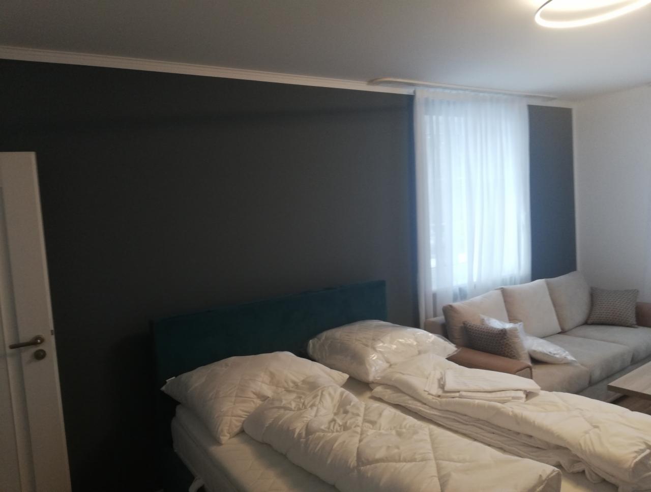 Pokój po odświeżeniu: ciemnoszara ściana z białą listwą przypodłogową, łóżko z zielonym zagłówkiem i białą pościelą w folii, beżowa sofa z poduszkami w tle, białe zasłony w oknie.