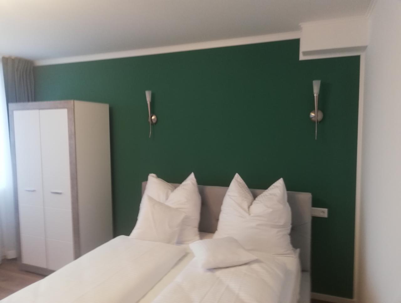 Sypialnia z łóżkiem dwuosobowym, zieloną ścianą i białymi lampkami ściennymi oraz szafą w kolorze białym i szarym.