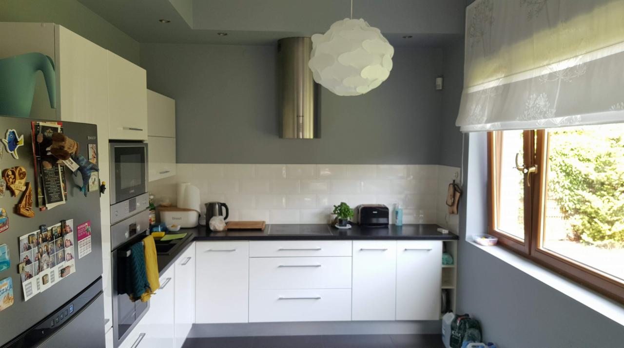 Nowoczesna kuchnia z białymi szafkami, czarnym blatem, białymi płytkami na ścianie, stalowym okapem i dekoracyjną lampą, widziana z perspektywy szerokokątnej.