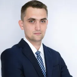 Portret młodego mężczyzny w granatowym garniturze i błękitno-białym krawacie, z poważnym spojrzeniem i krótkimi włosami, na jasnym tle.