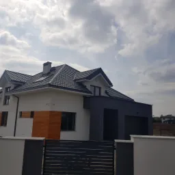 Nowoczesny dom jednorodzinny z szarą dachówką, biało-szarą elewacją z drewnianym akcentem, widok zza metalowej bramy wjazdowej w pochmurny dzień.