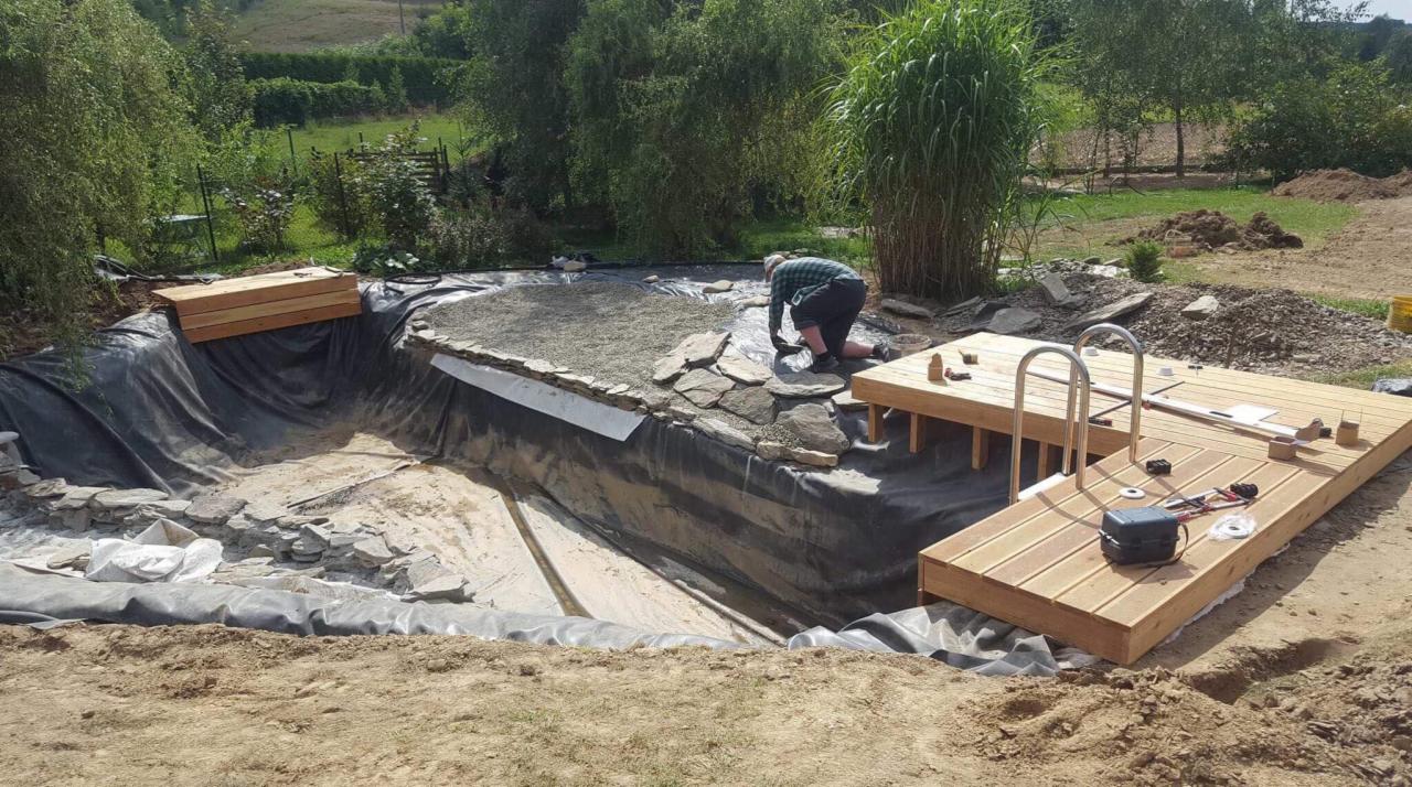 Budowa oczka wodnego z folią, kamieniami i drewnianym pomostem; pracownik na kolanach układa kamienie brzegowe, w tle trawa i drzewa.