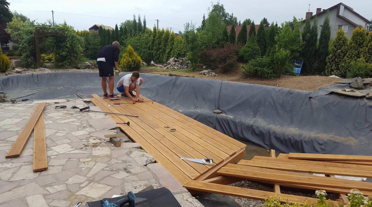 Budowa drewnianego tarasu nad stawem w ogrodzie, widoczni dwaj mężczyźni podczas montażu desek, w tle roślinność i dom.