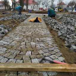 Robotnicy układają granitową kostkę brukową na placu budowy z widocznym kościołem w tle, używając łopat, młotka i drewnianej listwy do wyrównywania.