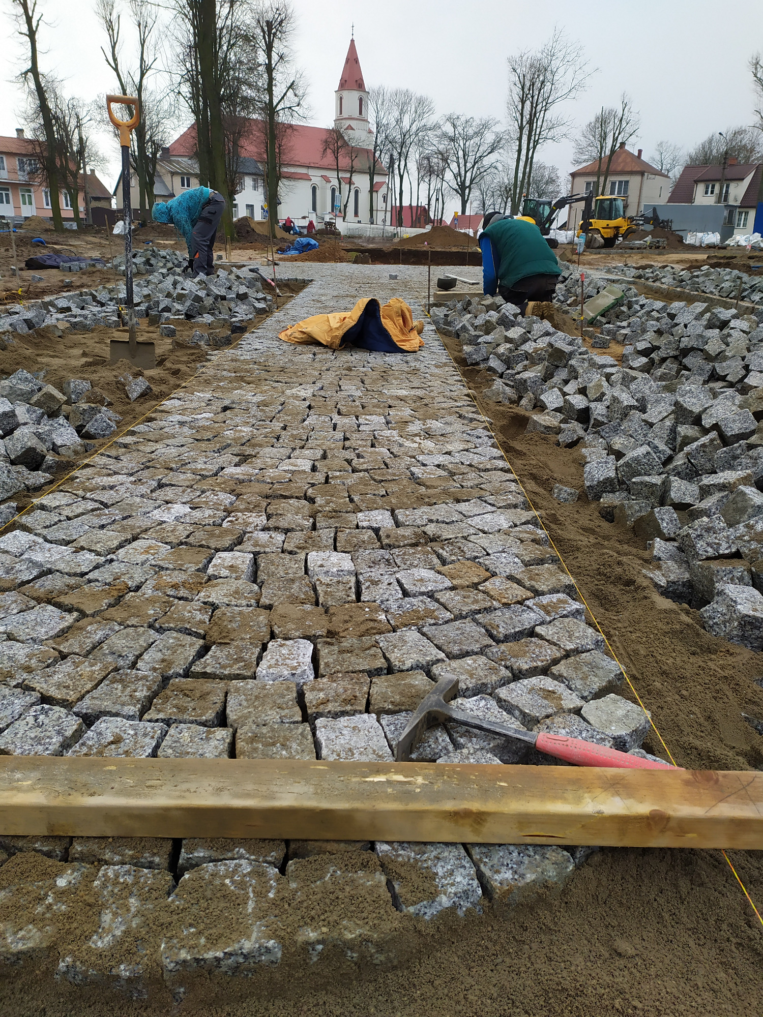 Robotnicy układają granitową kostkę brukową na placu budowy z widocznym kościołem w tle, używając łopat, młotka i drewnianej listwy do wyrównywania.