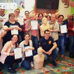 Grupa uśmiechniętych osób, w tym instruktor w koszulce ratownika medycznego, prezentuje certyfikaty ukończenia szkolenia z pierwszej pomocy, w tle manekiny do ćwiczeń resuscytacji i kolorowa ściana...