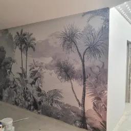 Ściana z fototapetą przedstawiającą motyw tropikalny w odcieniach szarości, widoczne narzędzia malarskie na podłodze, sufit z oświetleniem LED.