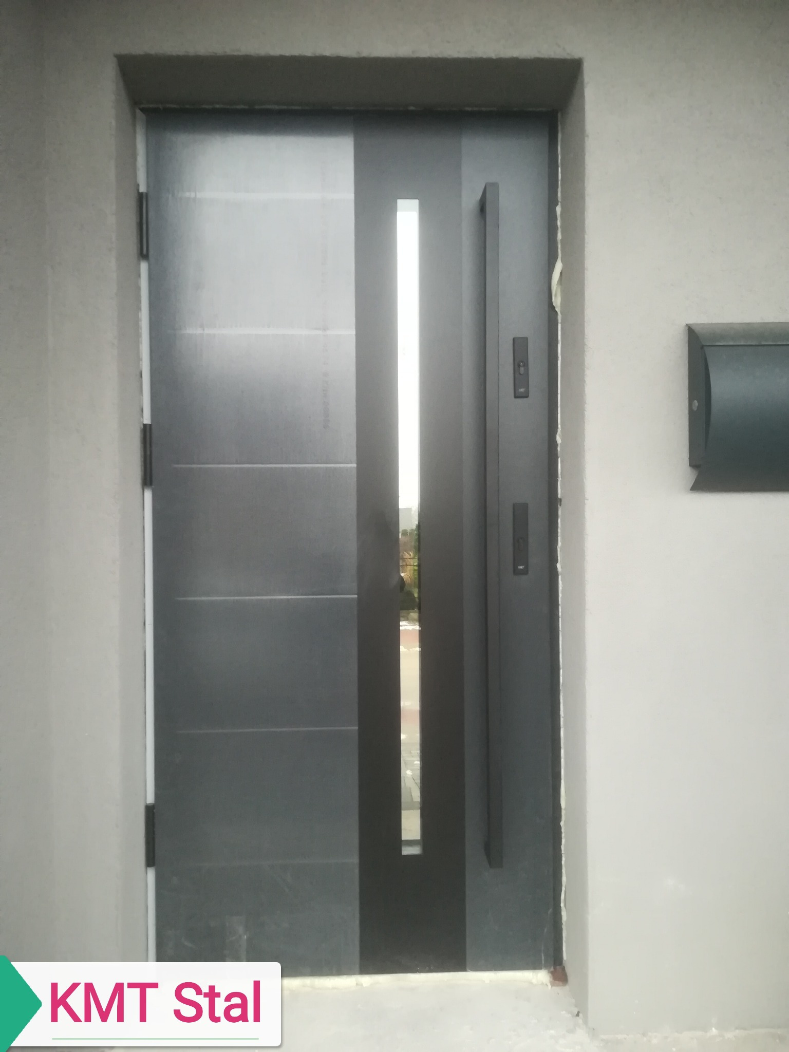 Nowoczesne, grafitowe drzwi wejściowe z długim uchwytem i wąskim przeszkleniem pionowym, zamontowane w szarej elewacji budynku z widoczną skrzynką na listy.