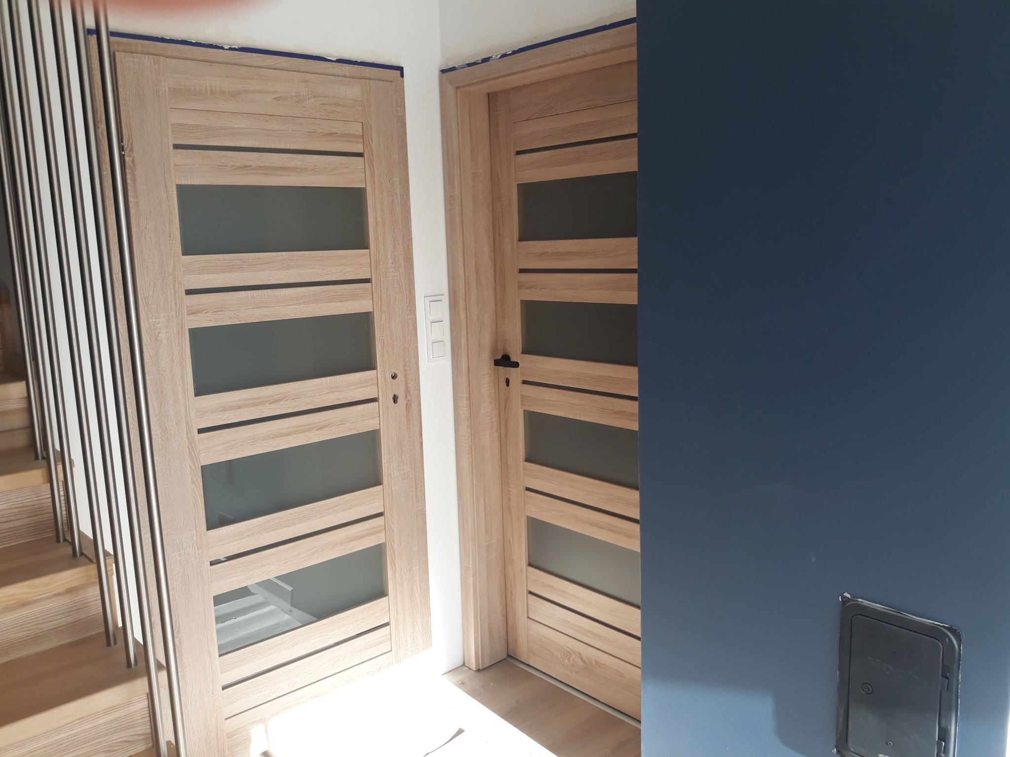 Dwie wewnętrzne drzwi z drewnianą okleiną i poziomymi przeszkleniami, zamontowane w nowym budynku, widoczne schody z metalową balustradą.