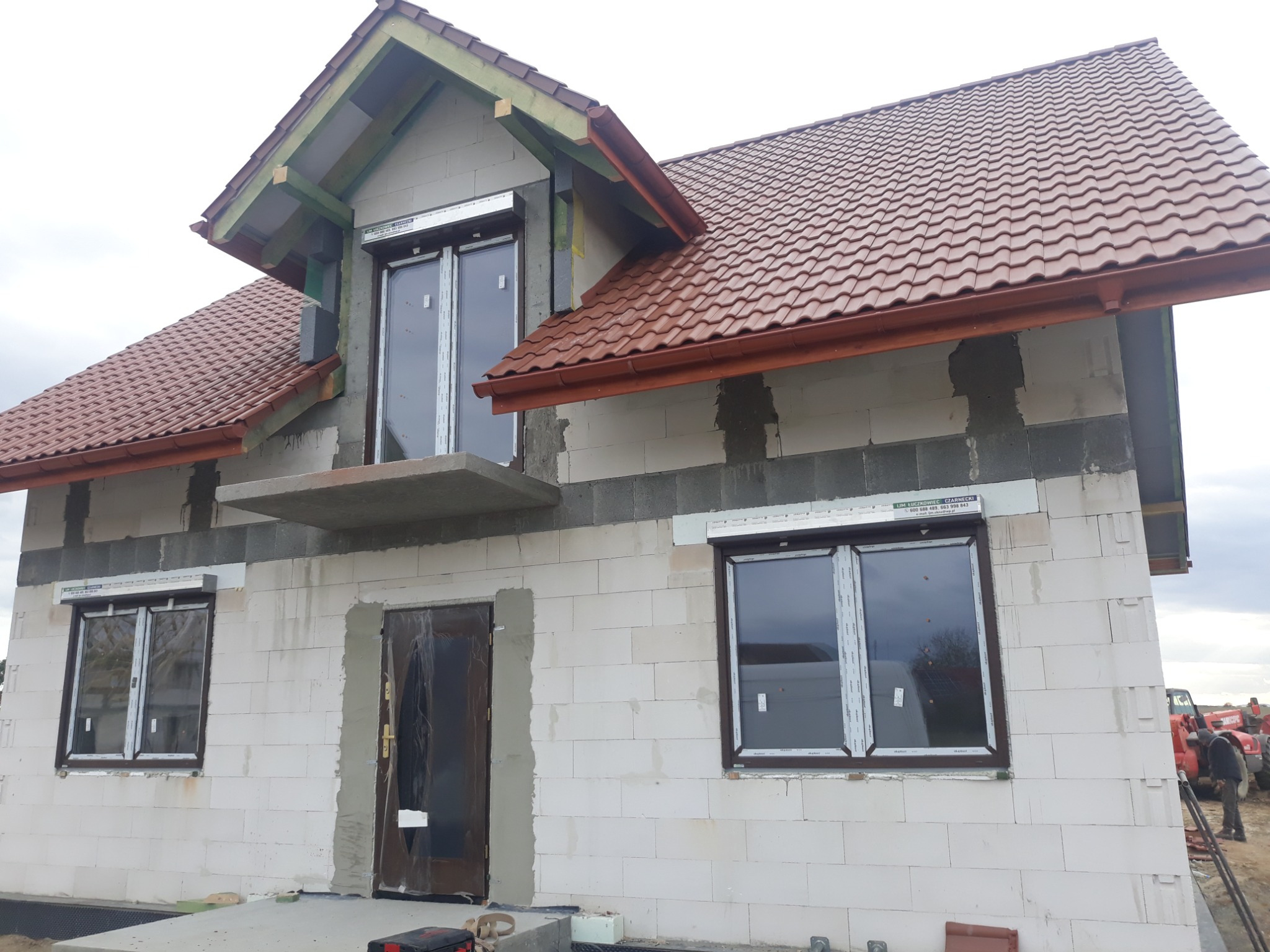 Niedokończony dom jednorodzinny z czerwoną dachówką, widoczne pustaki ceramiczne i zamontowane okna z roletami zewnętrznymi, na pierwszym planie skrzynka z narzędziami.