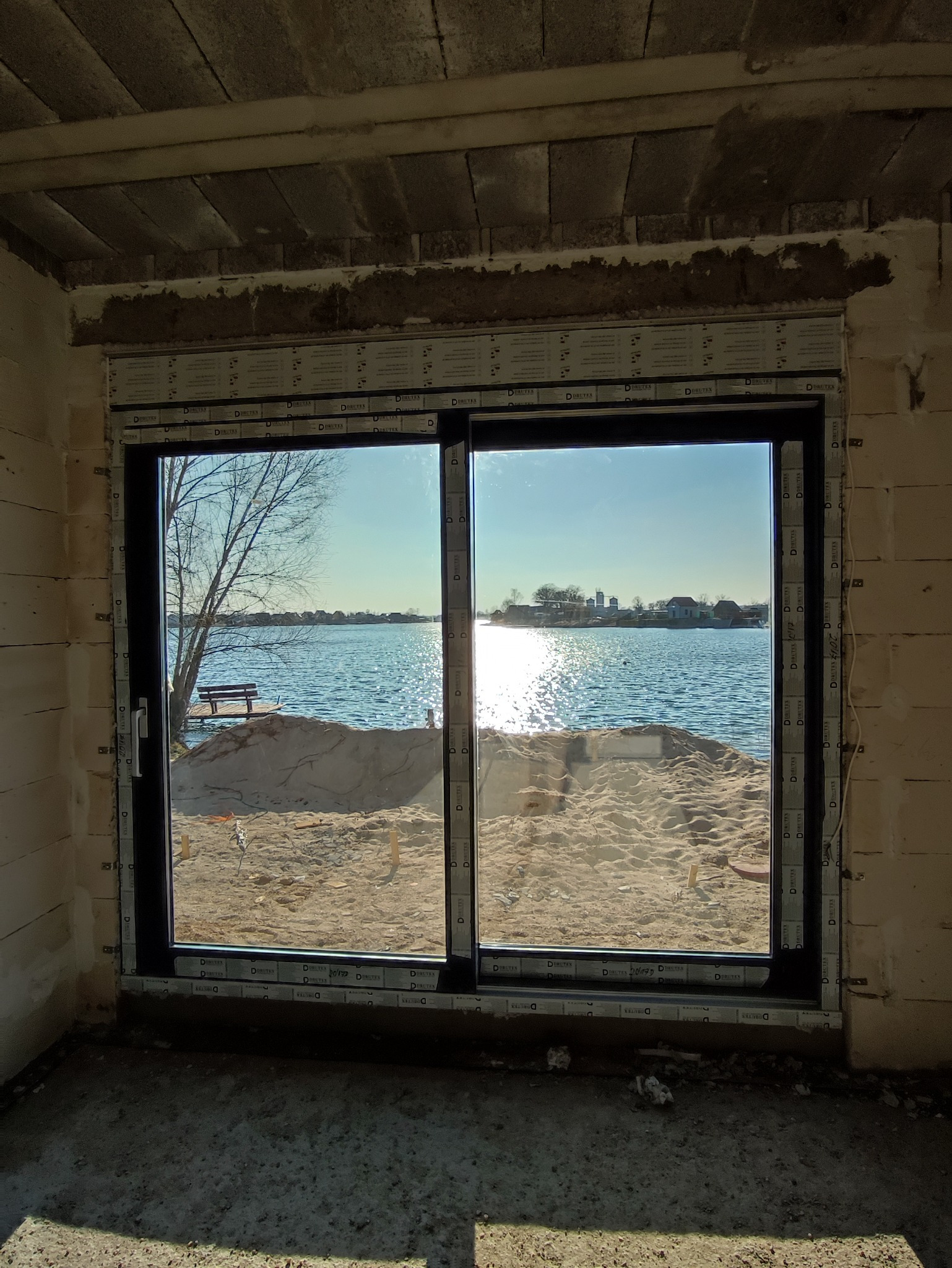 Nowe okno przesuwne z czarną ramą w surowym stanie, widok na jezioro z ławką na brzegu i odległe budynki, ślady prac budowlanych w pomieszczeniu.