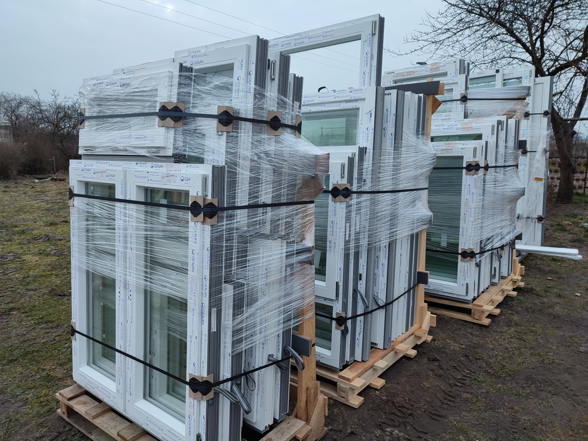 Palety z nowymi oknami PCV marki GEALAN, owinięte folią stretch i zabezpieczone taśmą, składowane na zewnątrz przed montażem.