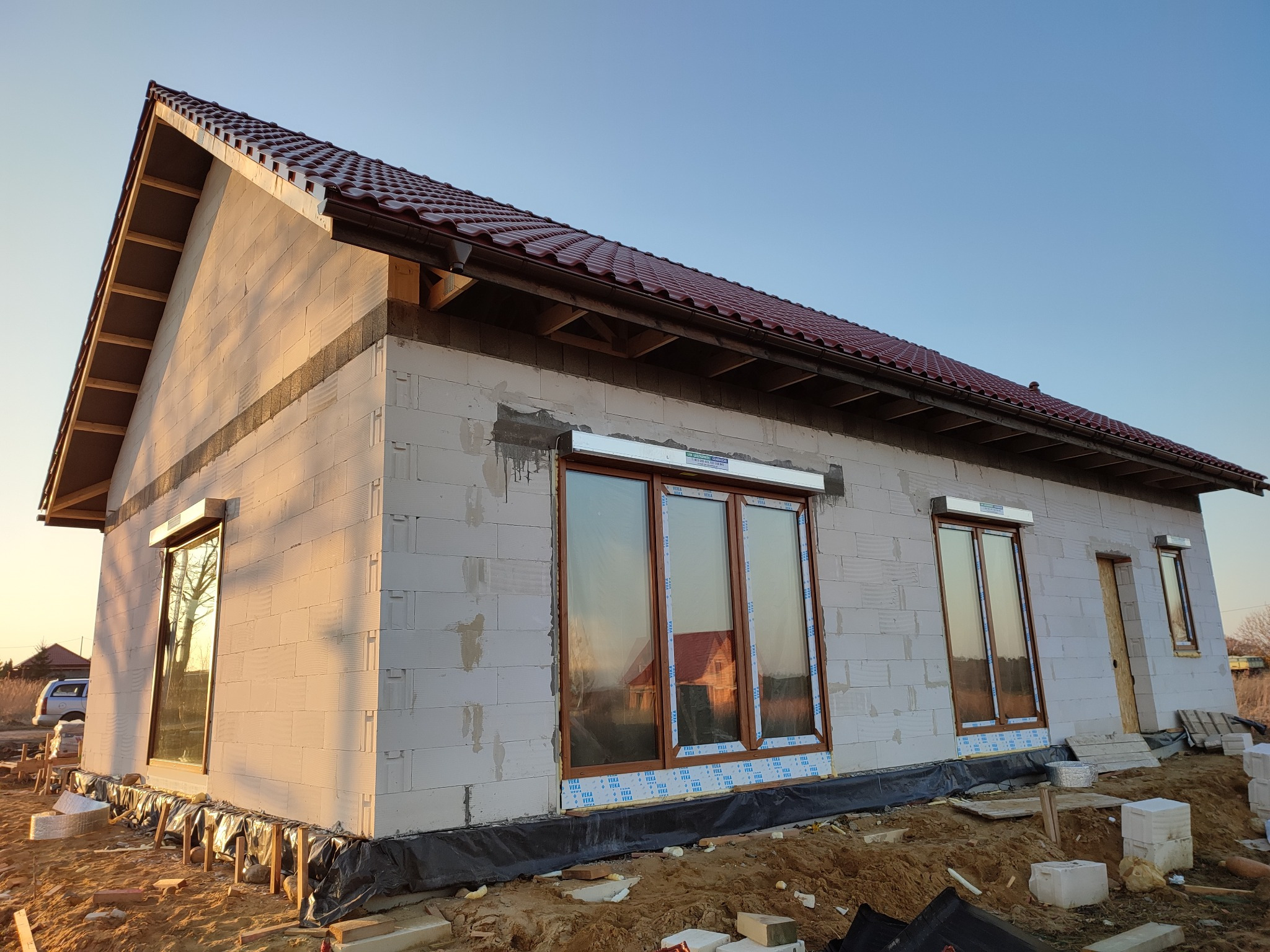 Niedokończony dom jednorodzinny z białego bloczka, z brązowym dachem i oknami z roletami zewnętrznymi, widoczne materiały budowlane i przygotowany teren.