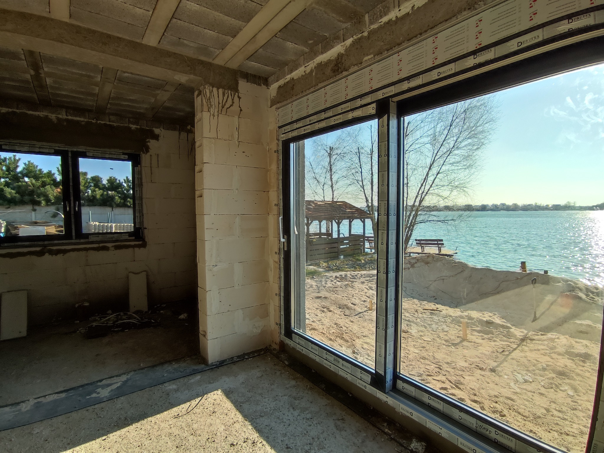 Wnętrze surowego budynku w trakcie budowy z widokiem na jezioro przez duże okno przesuwne z czarną ramą, w tle widoczna altana i pomost.