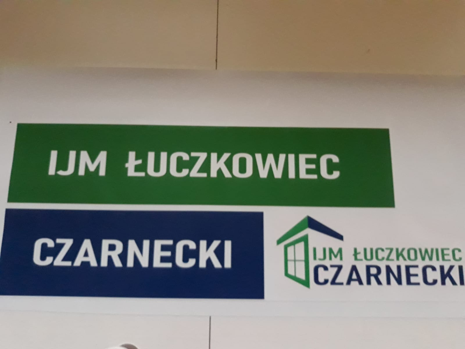 Tablica informacyjna z nazwami firm: IJM Łuczkowiec na zielonym tle i Czarnecki na granatowym tle, obok logo firmy zarys domu z oknem.