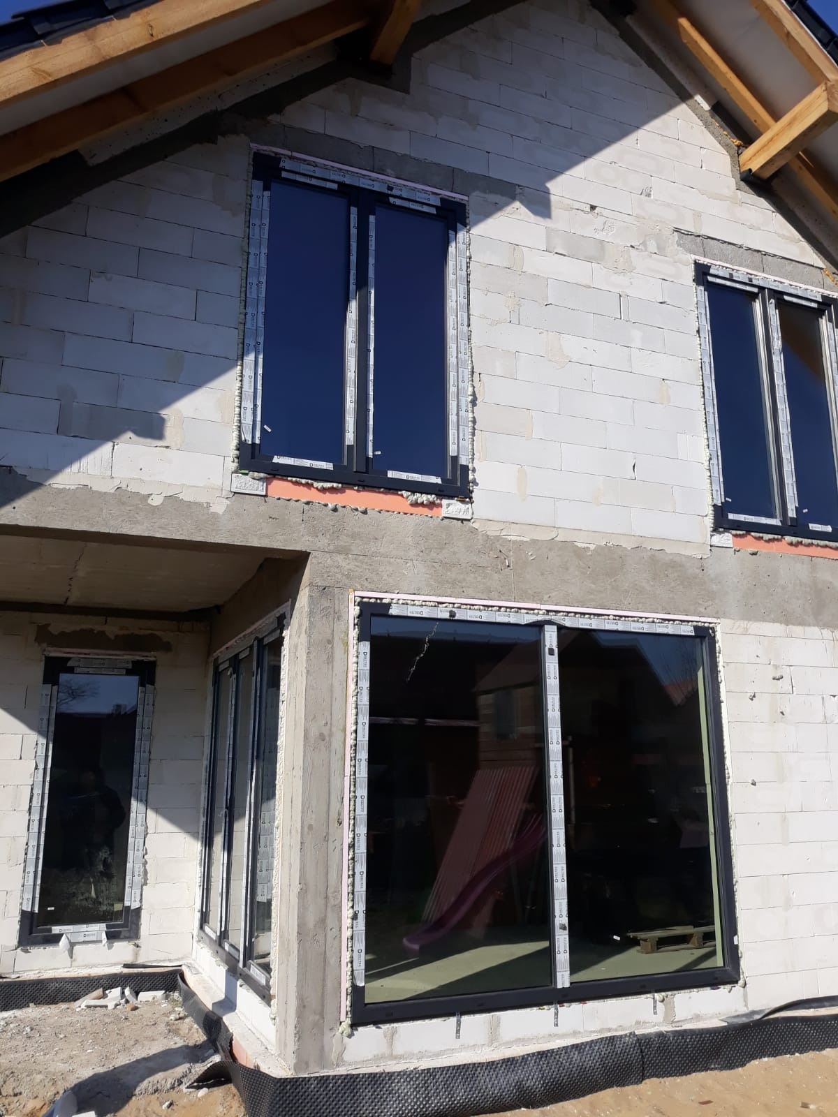 Surowy stan elewacji domu z zamontowanymi oknami w czarnych ramach, widoczne pianki montażowe i folia hydroizolacyjna przy fundamentach.