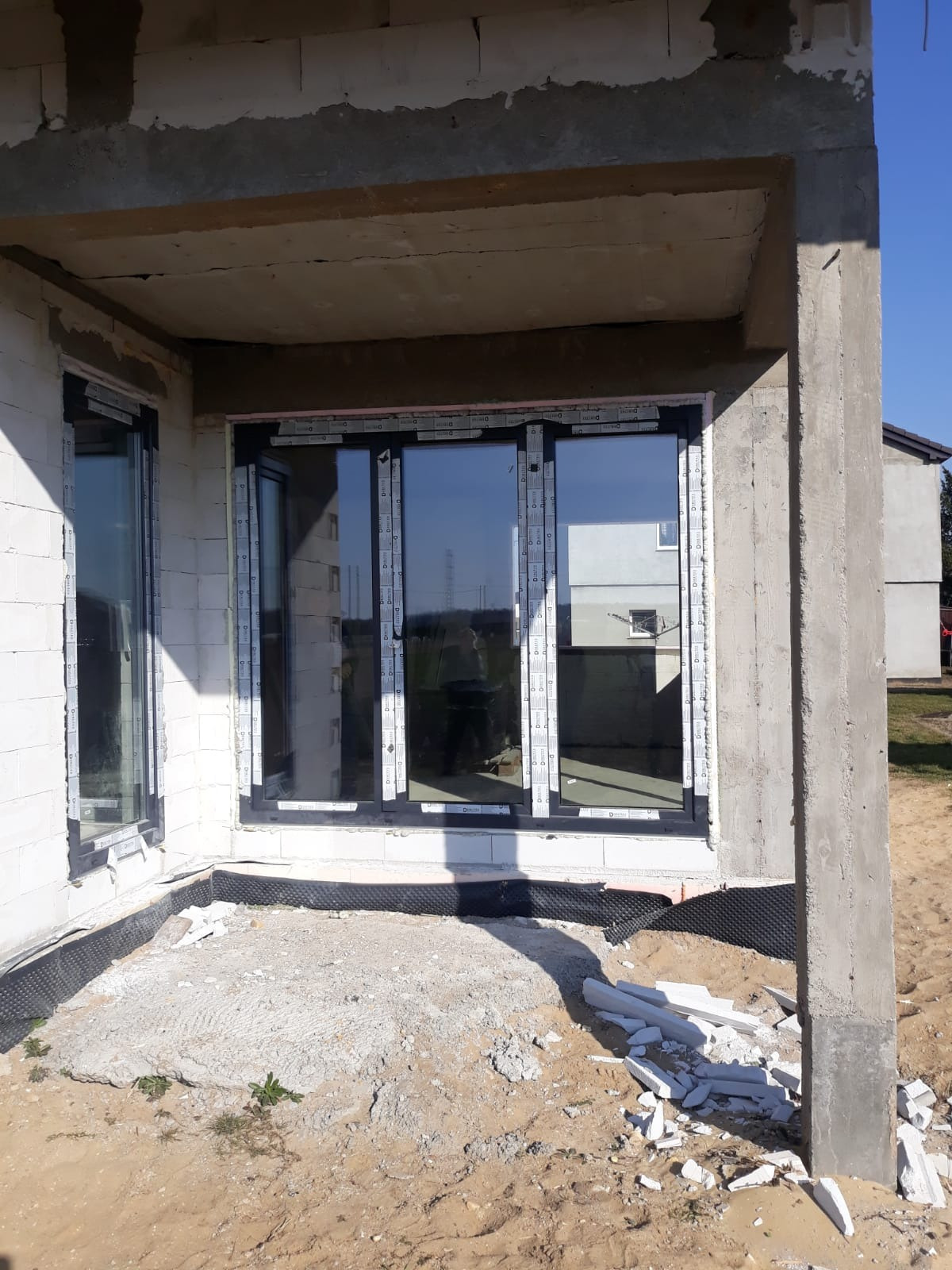 Surowy stan budynku z wstawionymi oknami PCV z czarnymi ramami, widoczne materiały budowlane i piasek na zewnątrz