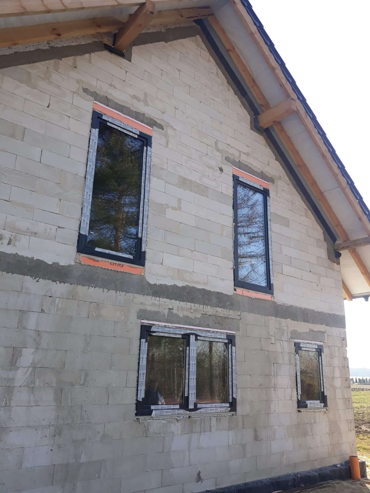 Surowy budynek z betonu komórkowego z zamontowanymi oknami z czarnymi ramami, zabezpieczonymi folią ochronną, widoczne drewniane elementy konstrukcji dachu.