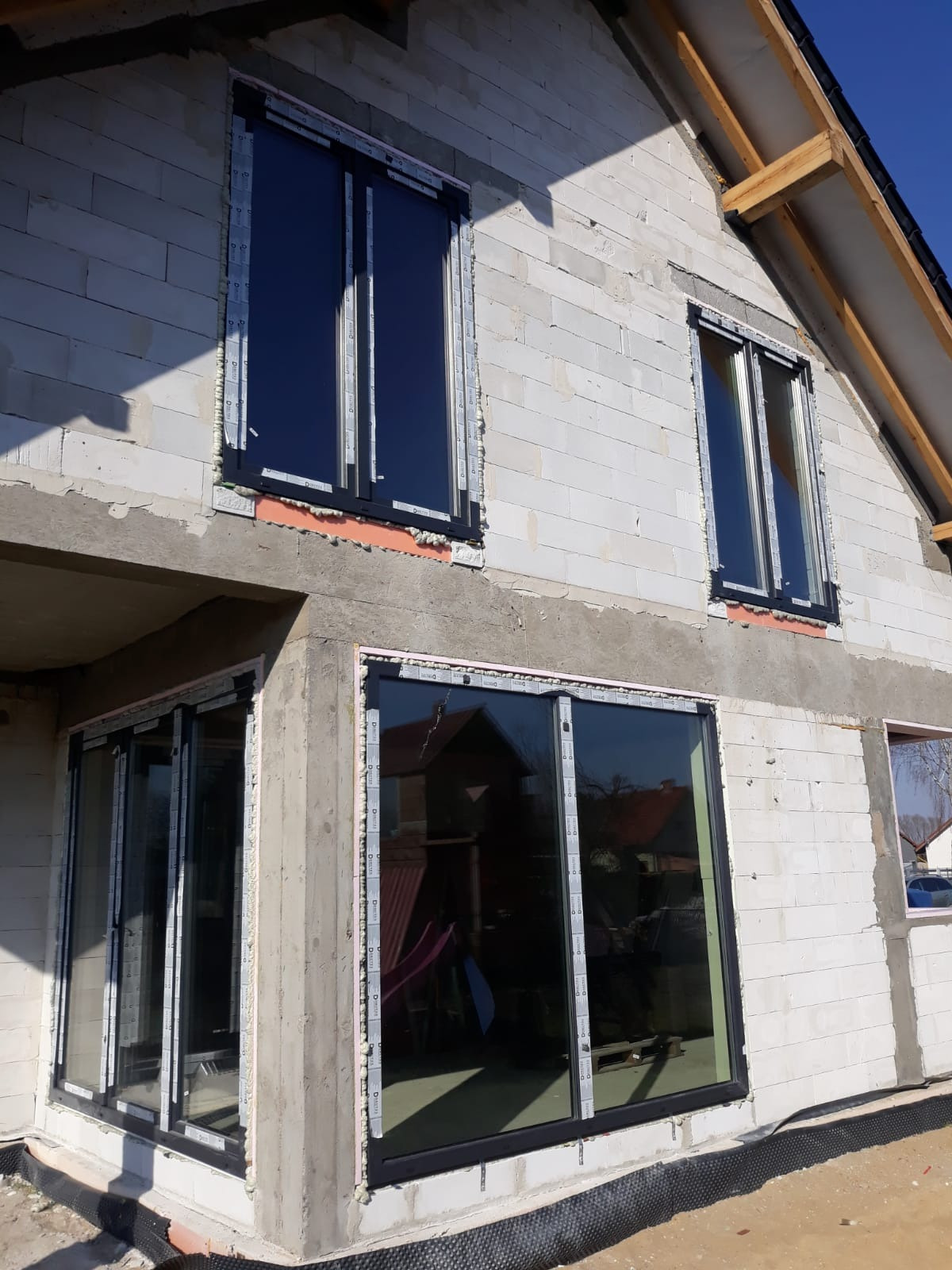 Surowy stan budynku z białego bloczka z zamontowanymi oknami w czarnych ramach, widoczne pianki montażowe i folie ochronne na profilach okiennych.