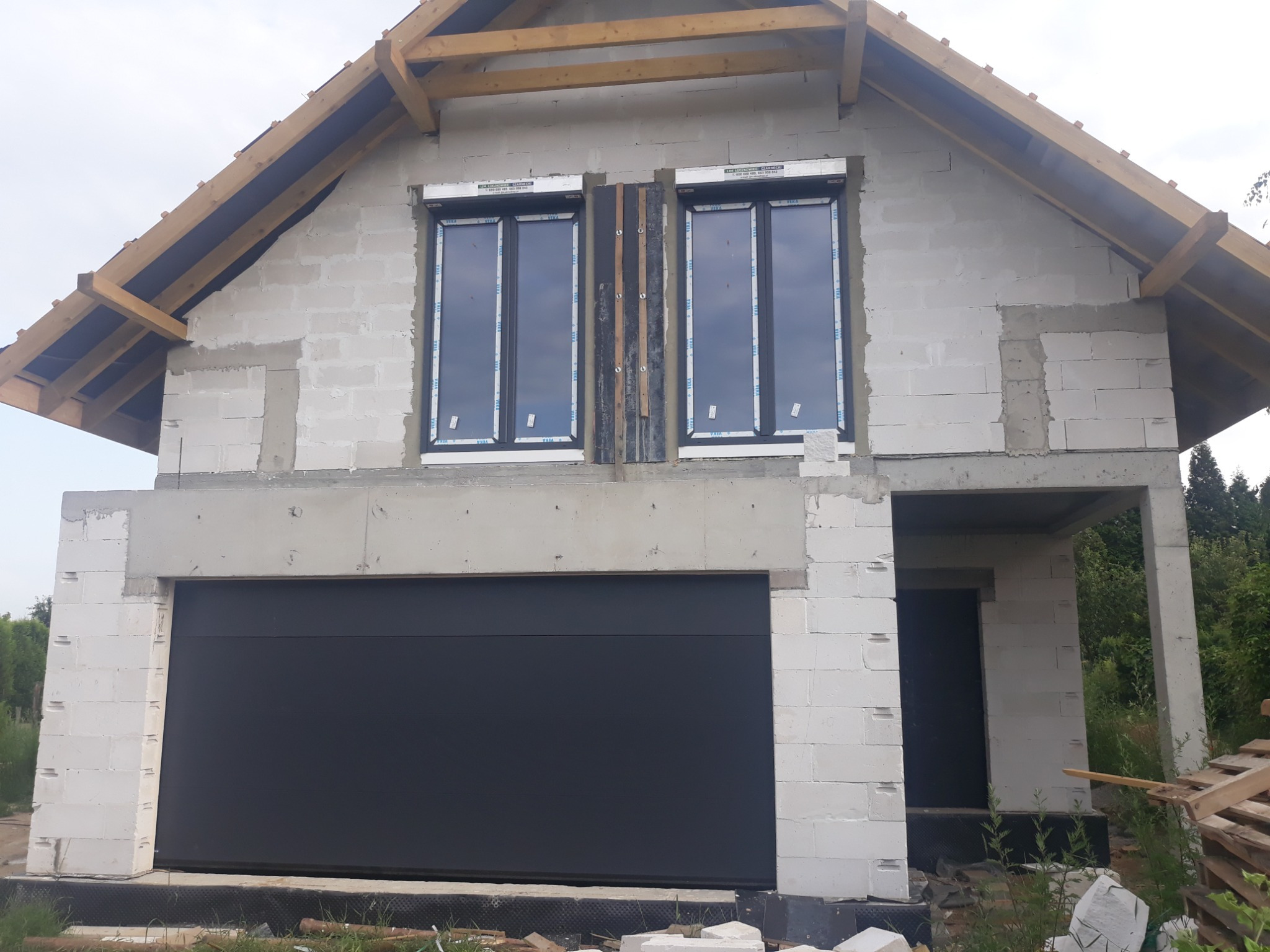 Surowy stan budowy domu jednorodzinnego z białych bloczków, widoczne okna z foliami ochronnymi i zamontowana brama garażowa, na tle nieba i drzew.