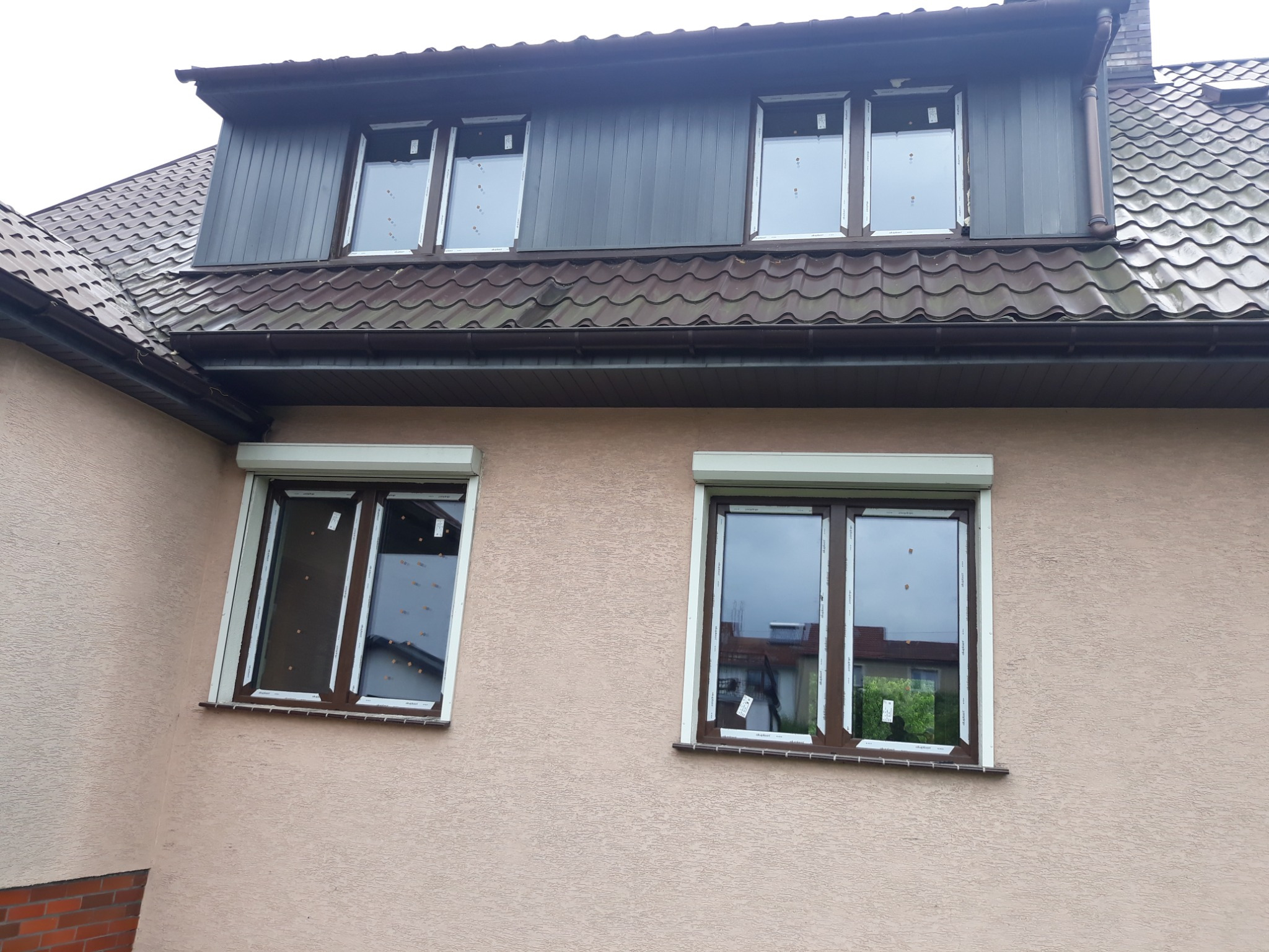 Elewacja budynku z oknami z roletami zewnętrznymi, widoczne ślady po montażu na szybach i ramach okiennych, brązowy dach z blachodachówki.