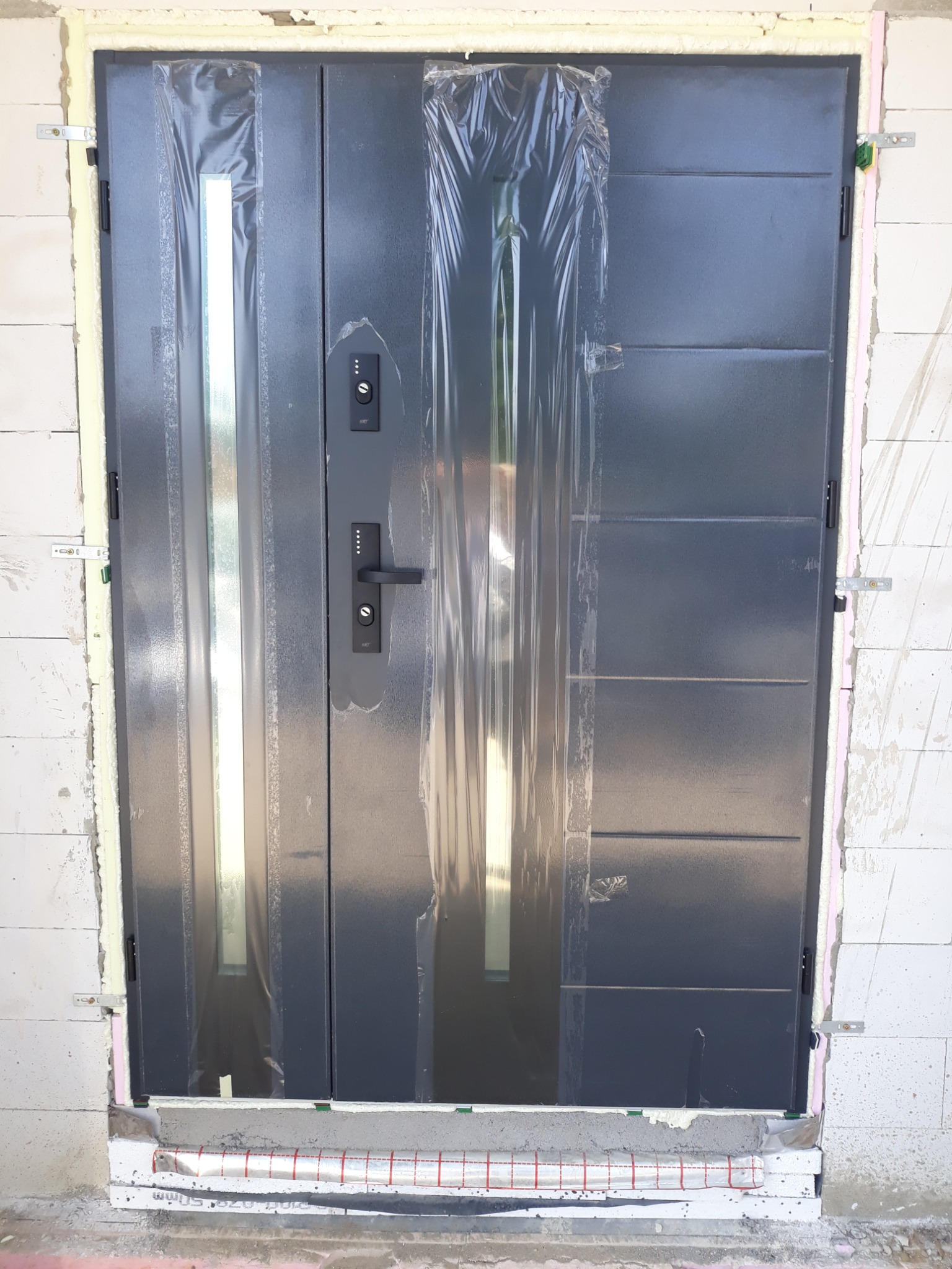 Nowe, szare drzwi wejściowe z długim pionowym przeszkleniem, częściowo zabezpieczone folią ochronną, zamontowane w surowym murze z widoczną izolacją.