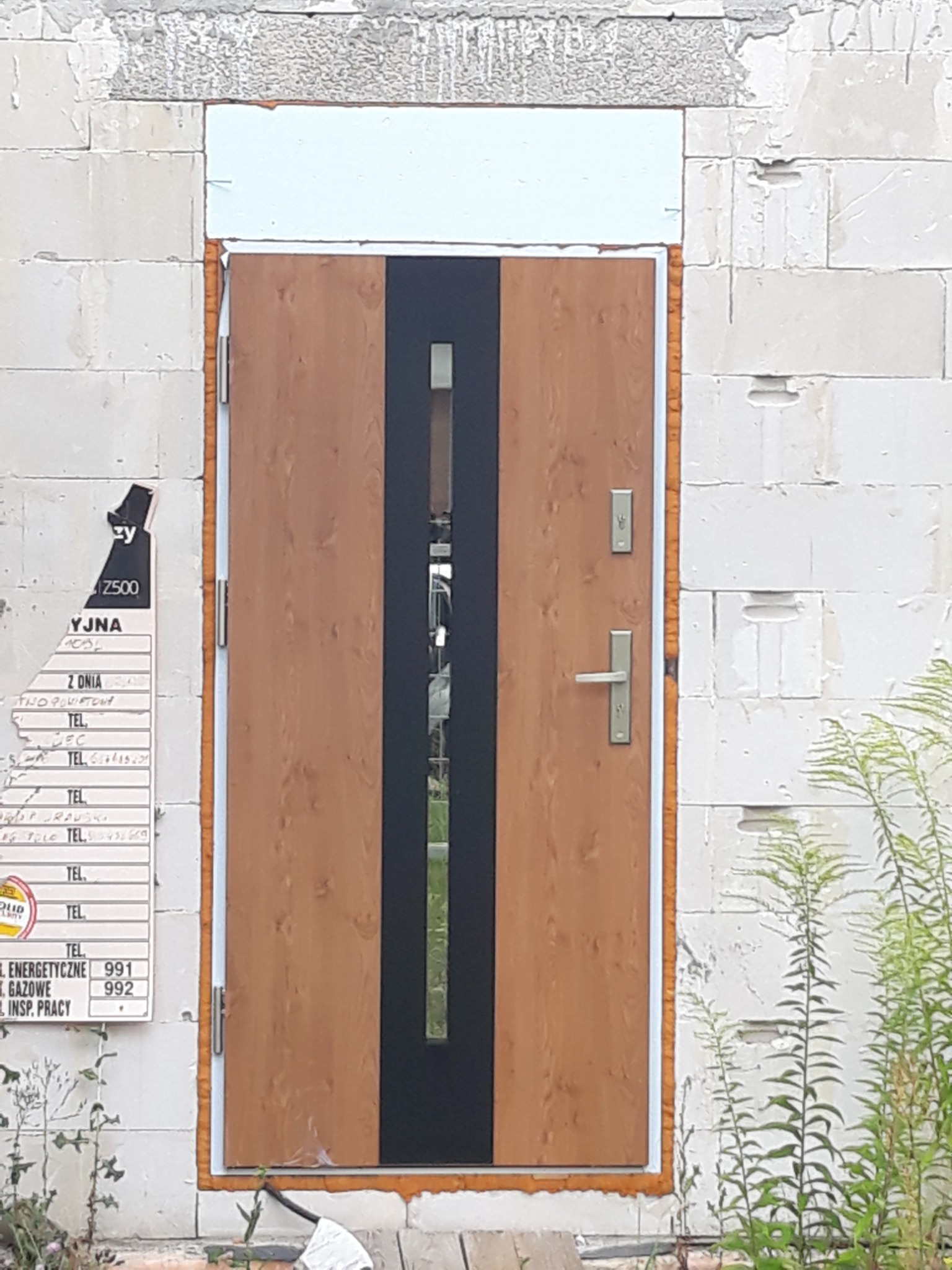 Brązowe drzwi wejściowe z czarnym pasem pośrodku, zamontowane w surowej ścianie z białych bloczków, z widoczną pianką montażową i ulotką informacyjną na ścianie.