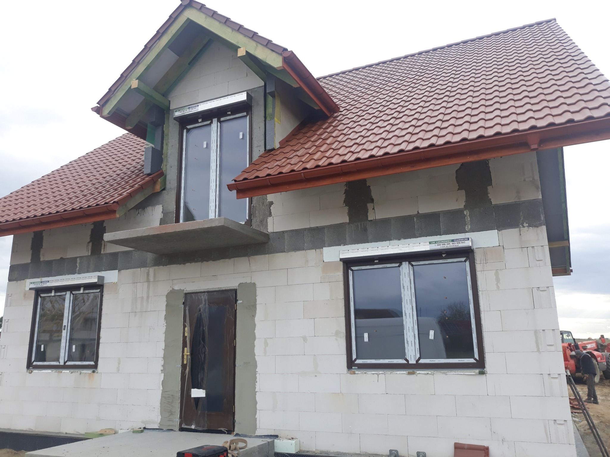 Surowy stan domu jednorodzinnego w trakcie budowy: białe bloczki, brązowa dachówka, zamontowane okna z roletami zewnętrznymi, widoczne elementy konstrukcji dachu.