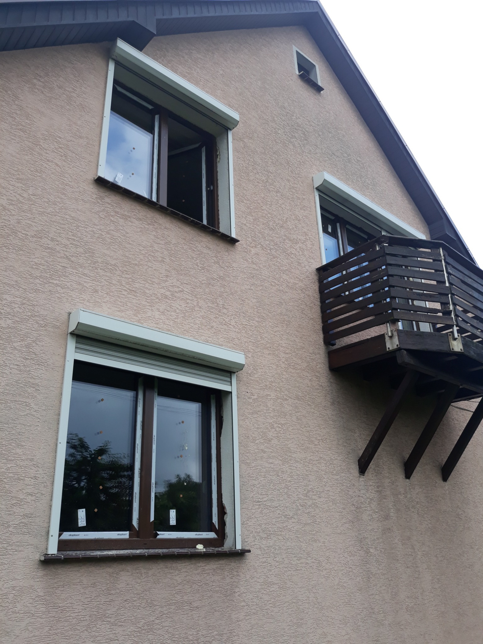 Elewacja domu z oknami z roletami zewnętrznymi, jeden z nich otwarty, oraz drewniany balkon z widoczną konstrukcją wsporczą.
