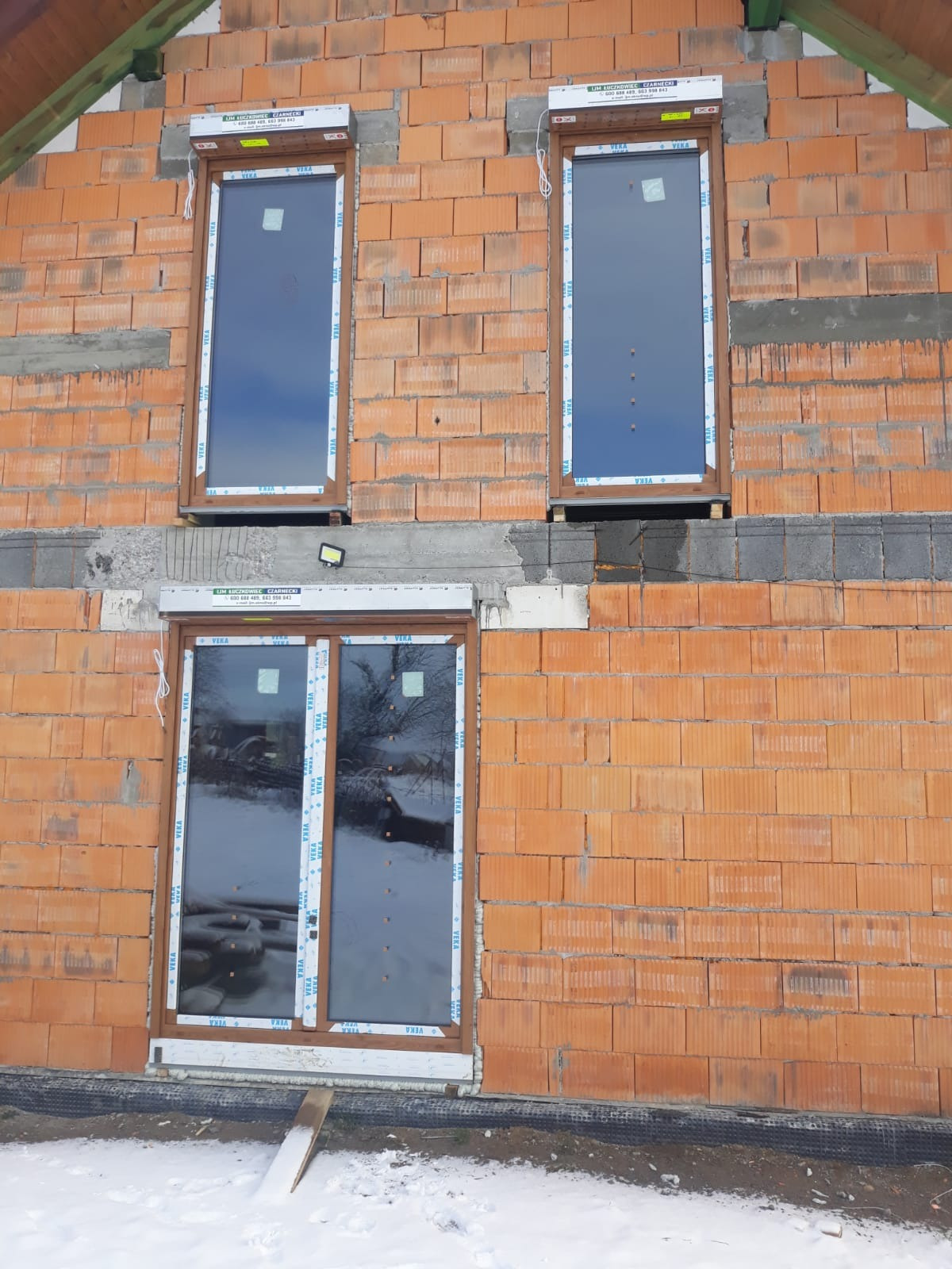 Montaż okien z brązowymi ramami w surowej ścianie z czerwonej cegły, widoczne rolety zewnętrzne i śnieg u podstawy budynku.