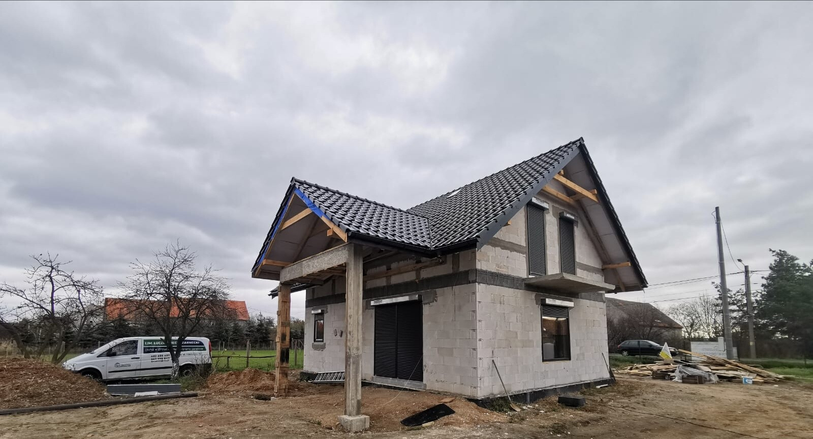 Surowy stan domu jednorodzinnego z czarną dachówką i szarymi roletami zewnętrznymi, budowanego z białych bloczków, widoczny samochód dostawczy z reklamą firmy w tle.
