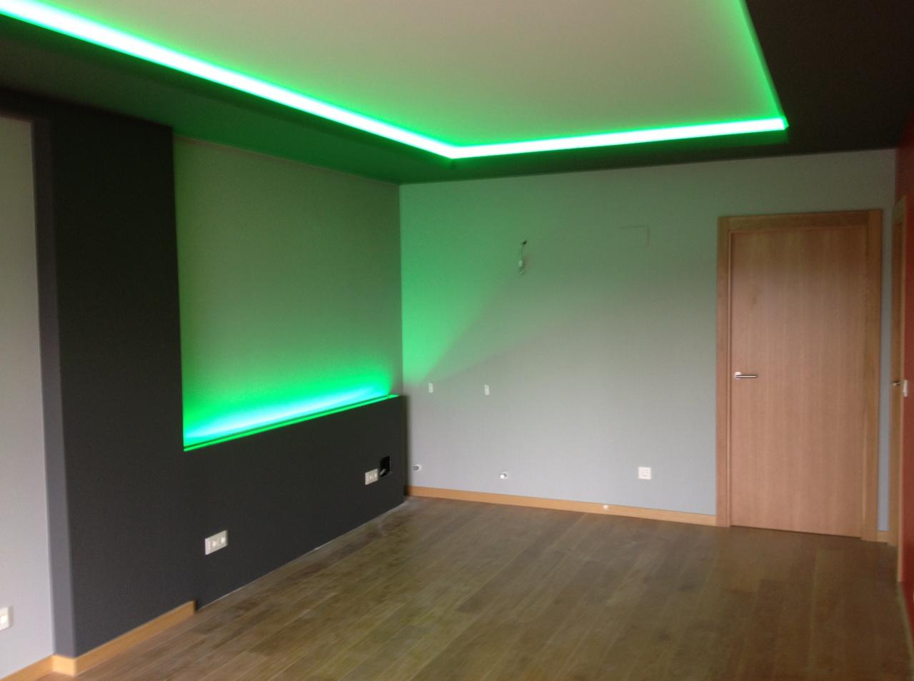 Pokój z podświetlanym sufitem i ścianą w kolorze zielonym neonu, drewniana podłoga, szare ściany, widoczne gniazdka elektryczne.