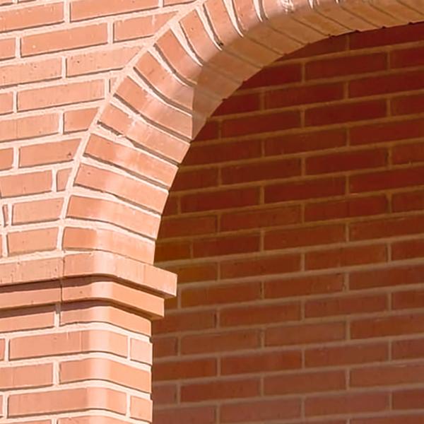 Fragment murowanej ściany z cegły w odcieniach czerwieni, tworzącej łuk nad wejściem, z widocznymi spoinami i detalami architektonicznymi.
