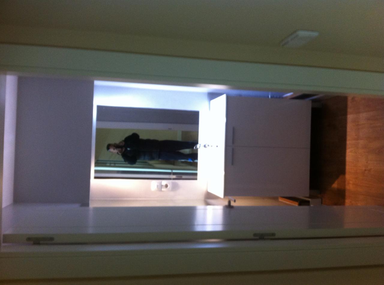 Nowoczesna kuchnia z białymi szafkami widziana z perspektywy blatu, odbicie osoby fotografującej w lustrzanej powierzchni nad blatem.