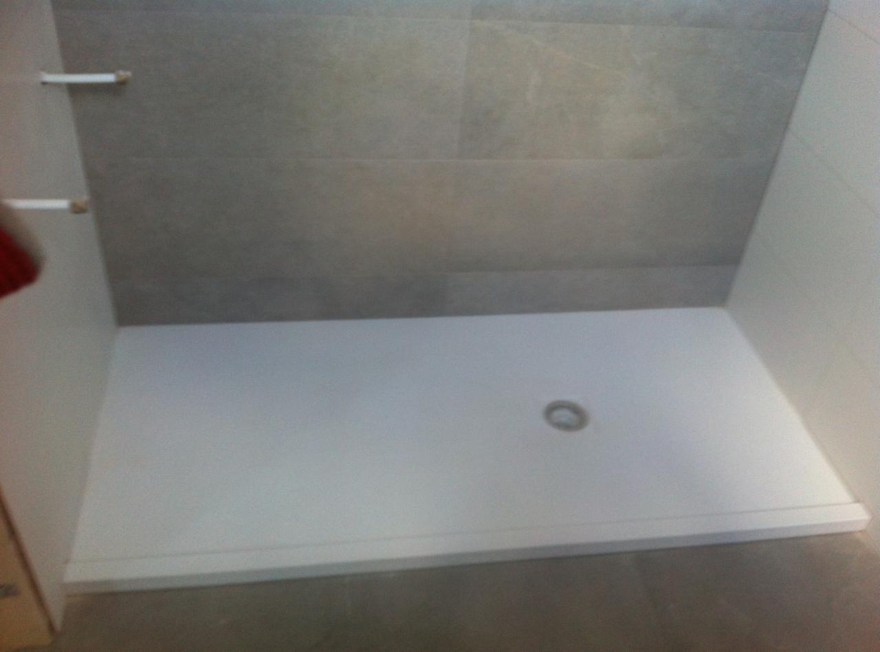 Biały brodzik z odpływem w rogu kabiny prysznicowej wyłożonej szarymi płytkami, widok z góry, minimalizm.