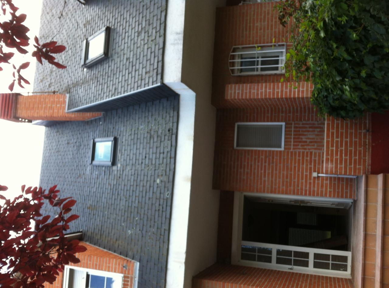 Fragment fasady budynku z cegły klinkierowej i szarej cegły, widoczne okna z kratami, zieleń i czerwone liście drzewa.