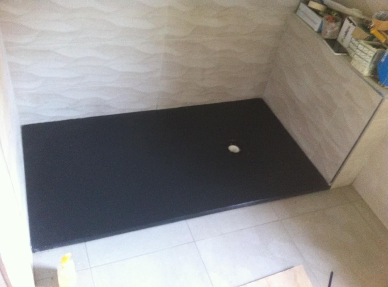 Nisko osadzony, czarny brodzik z odpływem w narożnej kabinie prysznicowej, wyłożonej beżowymi płytkami z falistym wzorem.