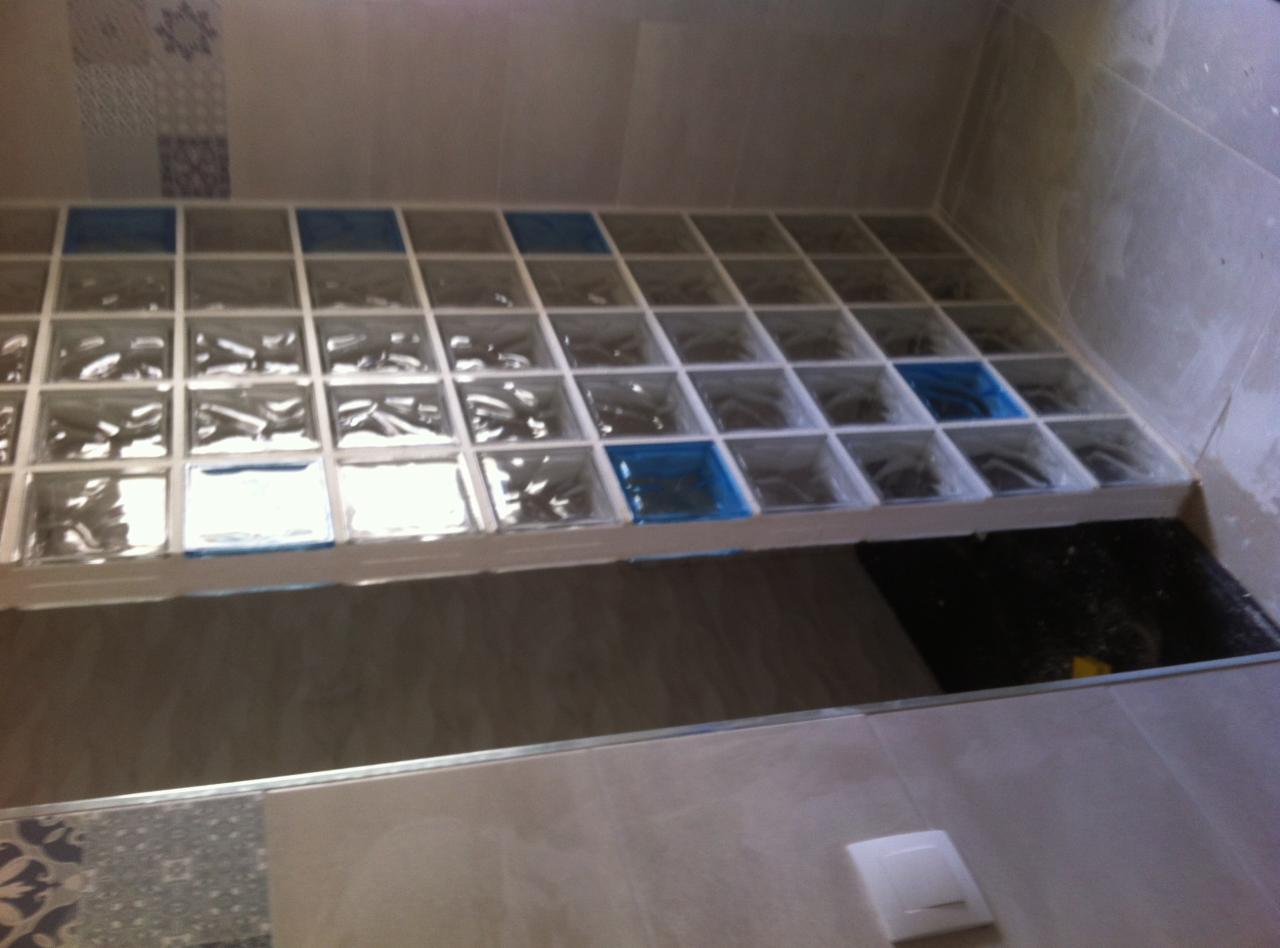 Montaż ścianki z luksferów w łazience, widoczne niebieskie zabezpieczenia na niektórych kostkach, jasne płytki na ścianach i podłodze, surowy stan podłoża pod ścianką.