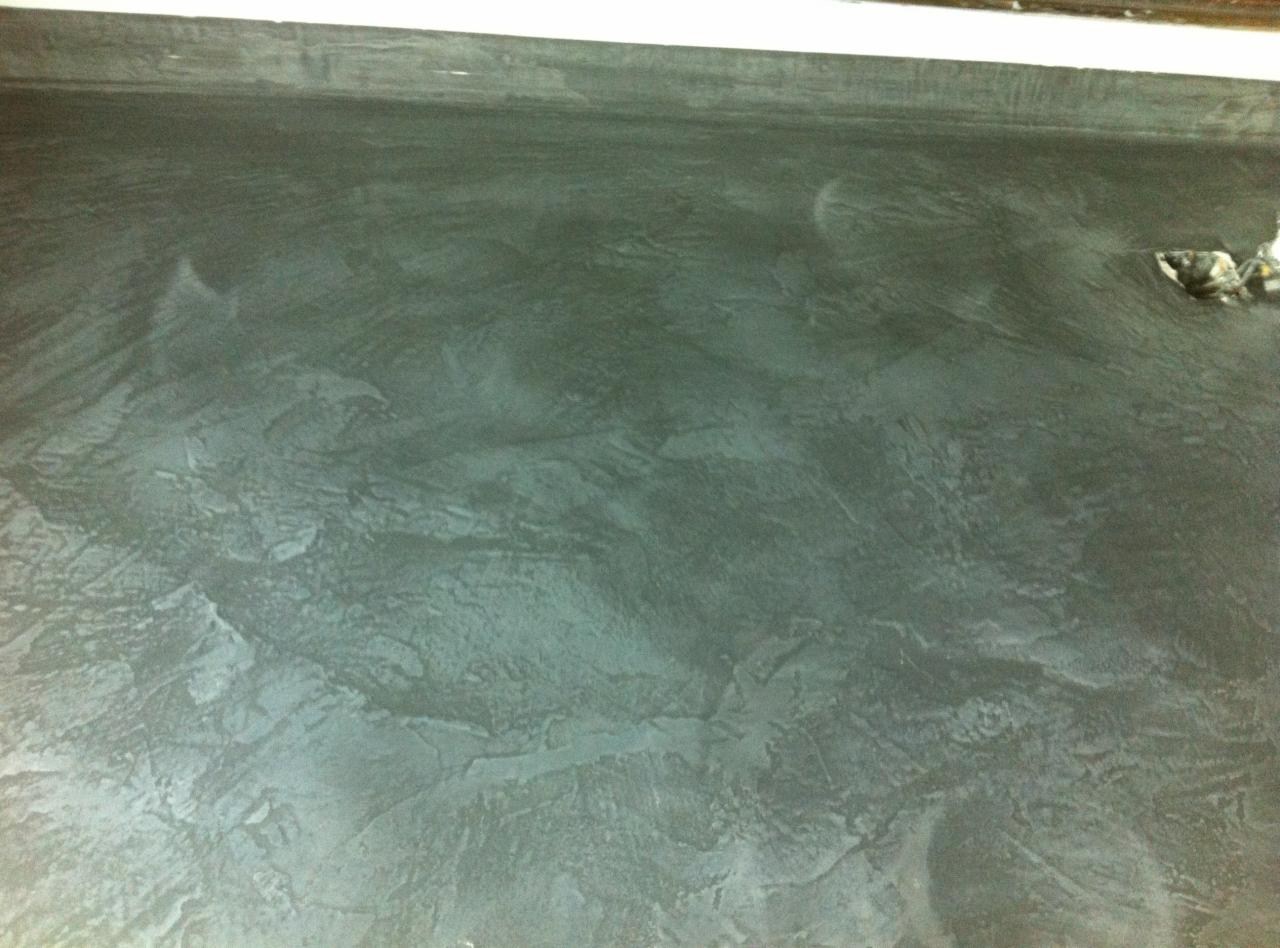 Szara, teksturowana podłoga z efektem betonu dekoracyjnego, widoczne ślady po pacy lub szpachelce, u góry biała listwa przypodłogowa, w prawym górnym rogu fragment puszki po farbie.