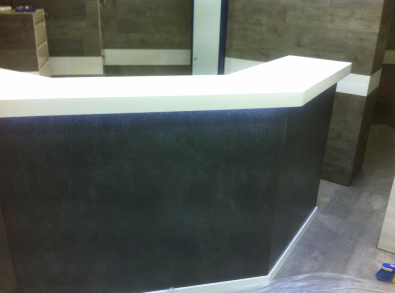 Nowoczesna lada recepcyjna z ciemną, teksturowaną okładziną i białym blatem, oświetlona od spodu, na tle ściany z paneli drewnopodobnych.