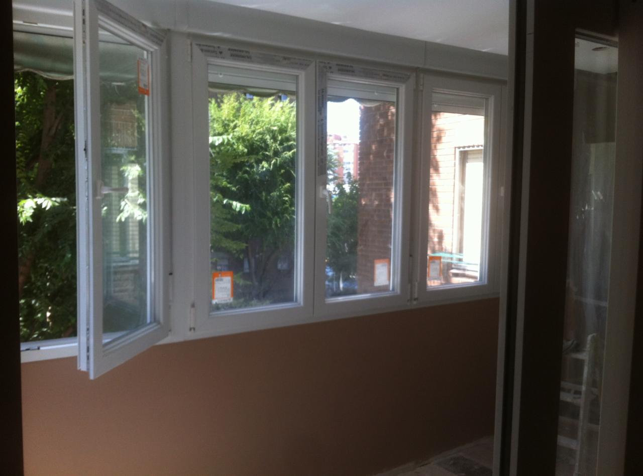 Zamontowane białe okna PCV na balkonie z widokiem na zieleń i budynek z czerwonej cegły. Jedno okno otwarte.